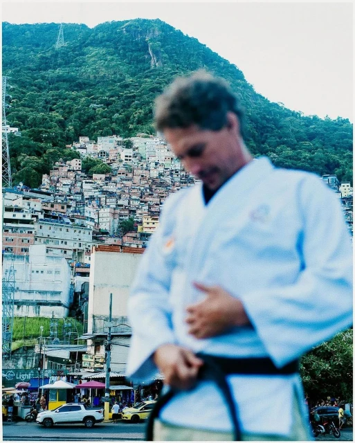 Man in a karate gi tying his belt, cityscape background, and mountain in background with vegetation in an outdoor daytime setting.