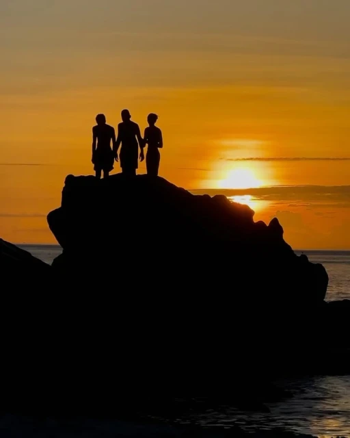 Silhouette of three people stand on a rocky cliff at sunset, with an orange sky reflecting on the ocean's surface below.