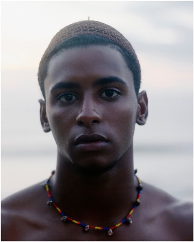 Close-up portrait of a young adult male with dark skin, a knitted cap, and a beaded necklace, posing outdoors with a serene expression.