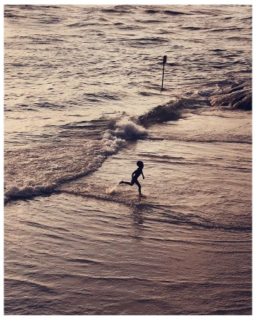 Silhouette of a child running along a sandy beach during golden hour with gentle waves and a marker pole in the background.