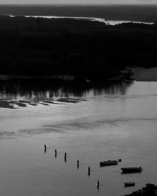 Monochrome high-angle shot features silhouetted people standing in water, small boats, and background trees and distant land.