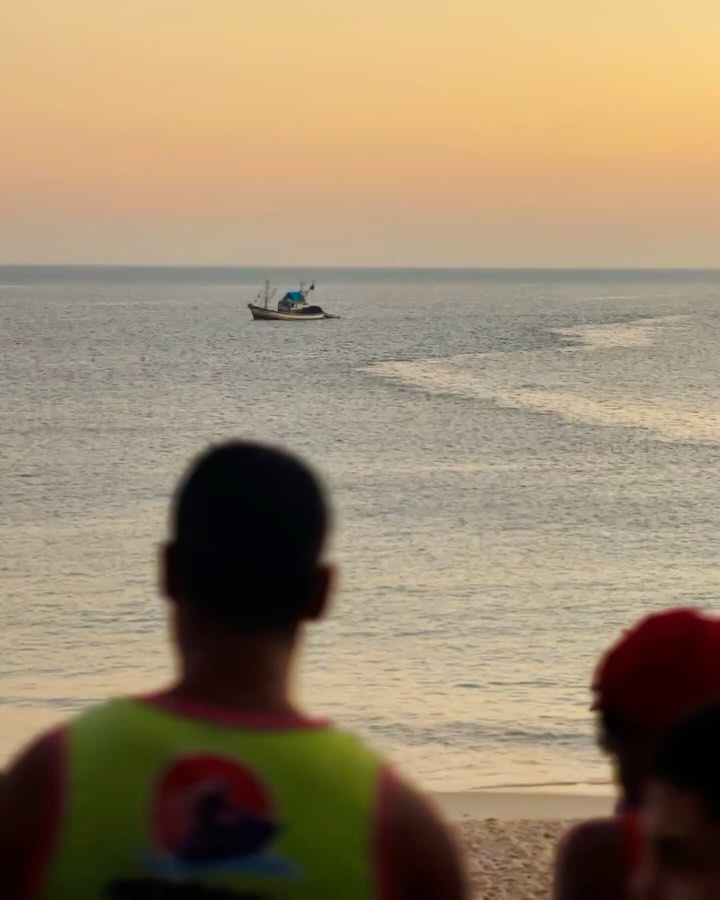 People look out at a fishing boat on the water under a pale orange sky at sunset from the beach.