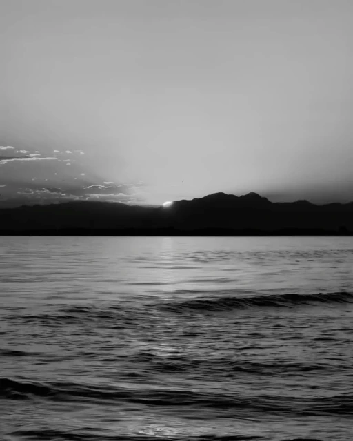 Black and white seascape featuring a calm ocean reflecting the setting sun, with silhouetted mountains in the distance and scattered clouds.