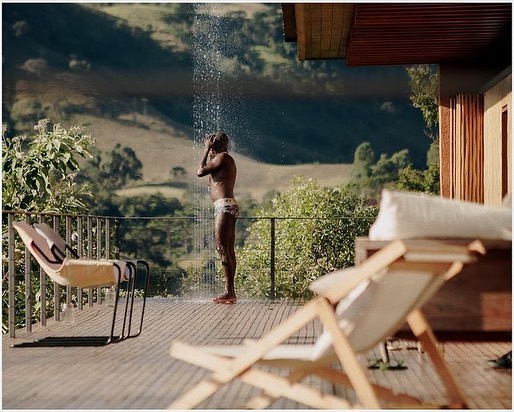 Adult male showering outdoors on a wooden deck with green hills and vegetation in the background, captured on a sunny day.
