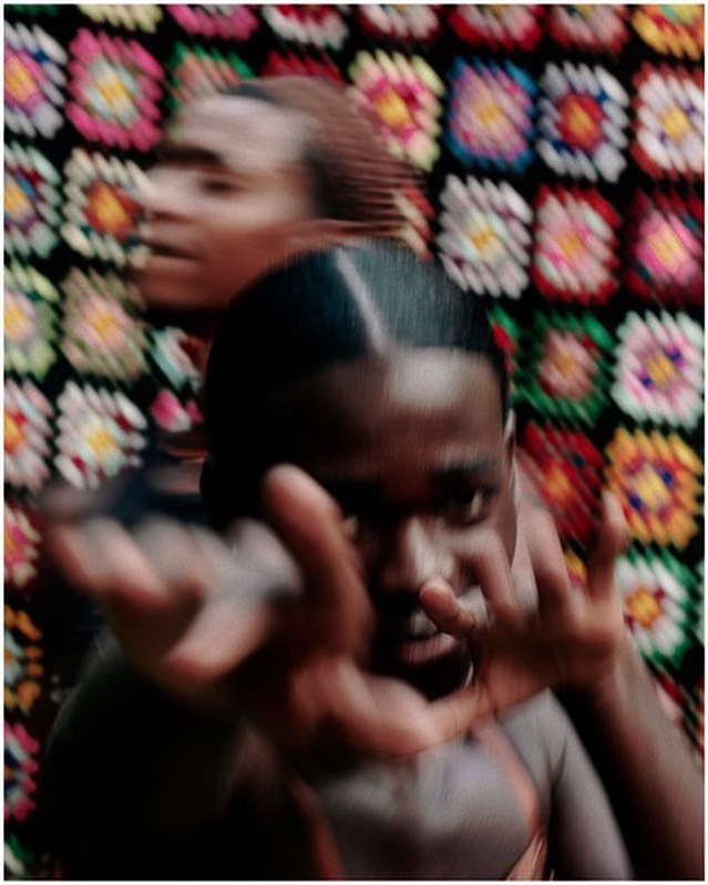 Two young Black adults in motion against a colorful, patterned textile backdrop, creating a dynamic, blurred effect in an indoor setting.