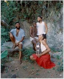 Four Black men posing outdoors in a lush natural setting with rocks and foliage, accompanied by a cat.