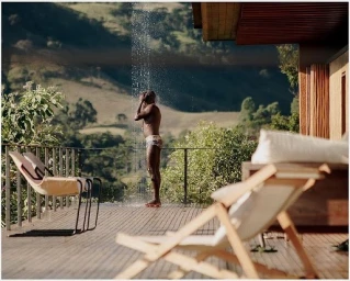 Man showering under an outdoor shower on a wooden deck overlooking a mountain valley, photographed by Rodrigo Oliveira.
