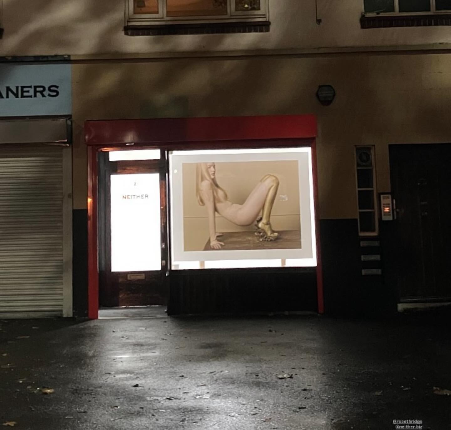 An advertising poster in a store window features a slender, fair-skinned woman in gold heels posing against a neutral background at night.