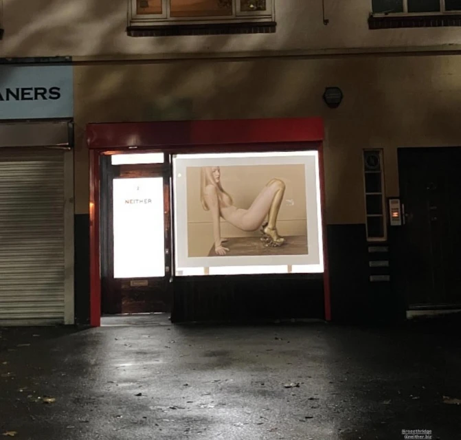 An advertising poster in a store window features a slender, fair-skinned woman in gold heels posing against a neutral background at night.