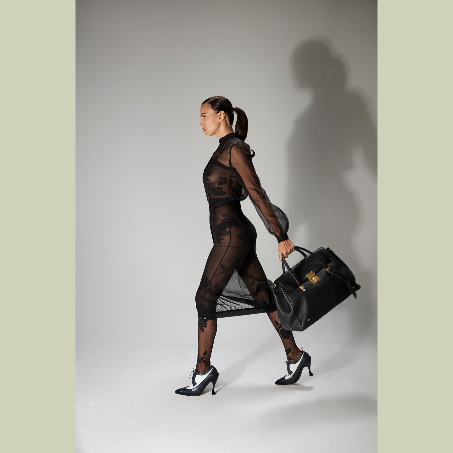A woman in a sheer black dress walks with a black leather handbag against a neutral background in a studio fashion shot.
