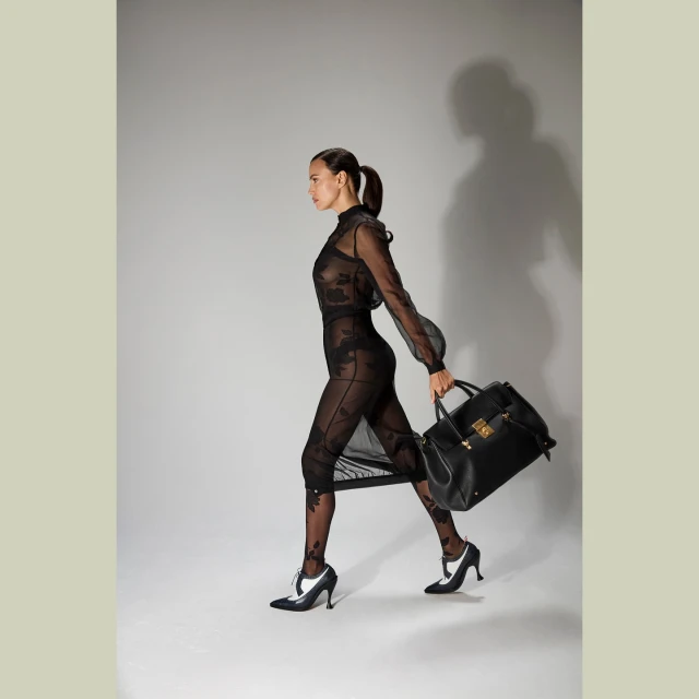 A woman in a sheer black dress walks with a black leather handbag against a neutral background in a studio fashion shot.