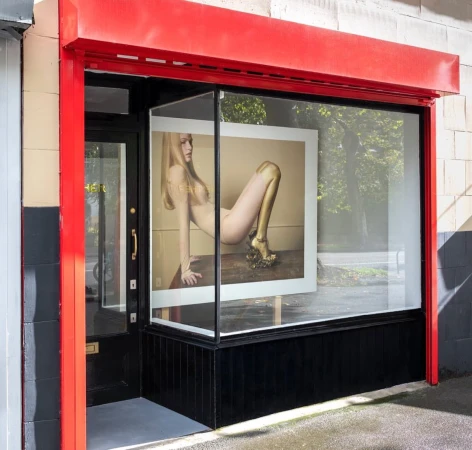 Fashion photo of a topless woman with golden painted skin and golden shoes inside a storefront window with a red awning.