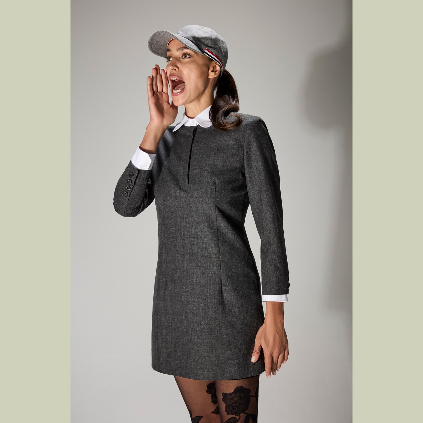 Young woman in a grey dress, cap, and patterned tights, shouting with her mouth open, set against a neutral studio backdrop. Fashionable and energetic.