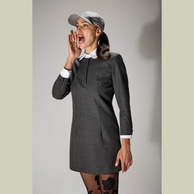 Young woman in a grey dress, cap, and patterned tights, shouting with her mouth open, set against a neutral studio backdrop. Fashionable and energetic.