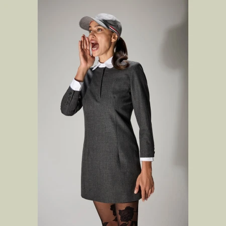 Young woman in a grey dress, cap, and patterned tights, shouting with her mouth open, set against a neutral studio backdrop. Fashionable and energetic.