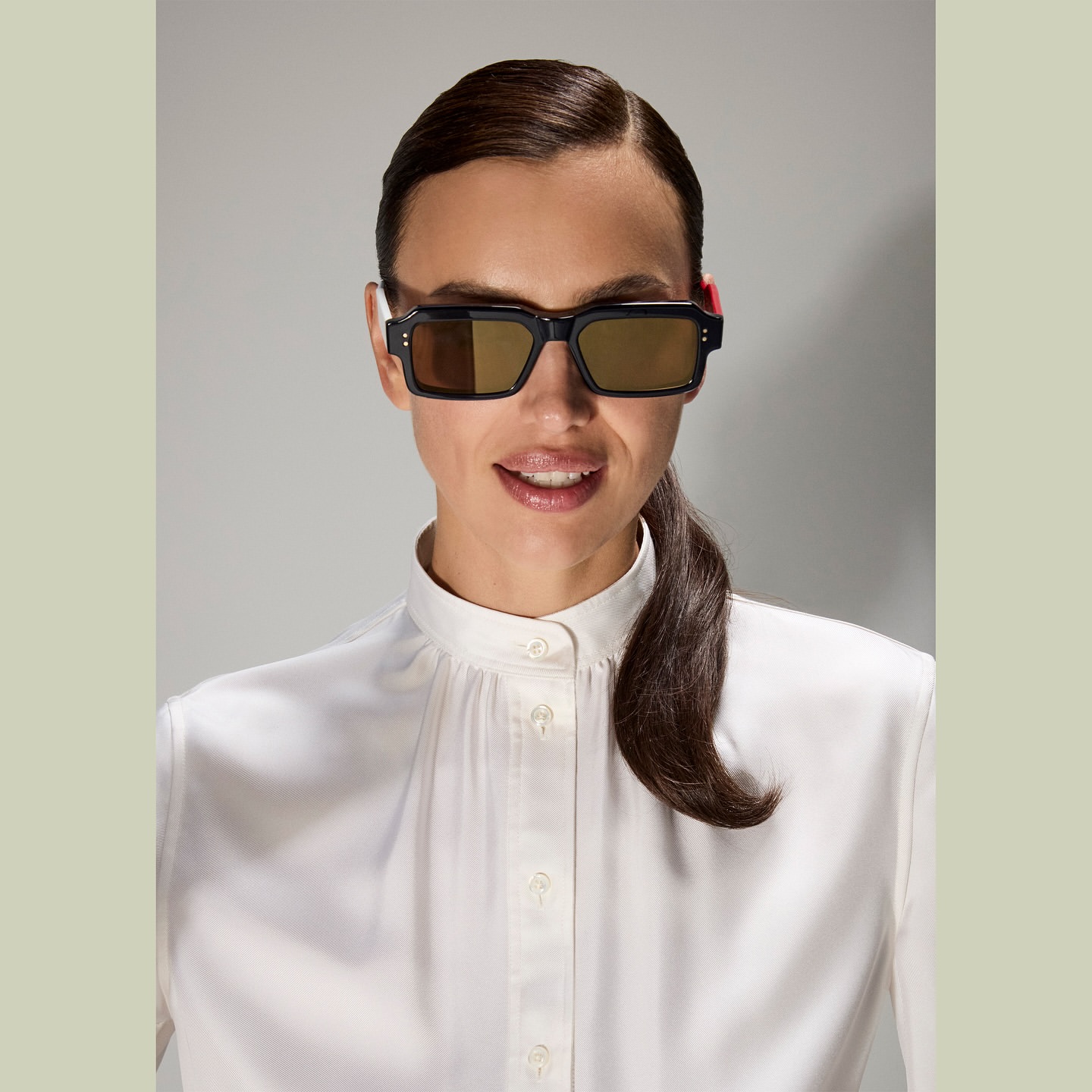 A woman in a white blouse and black sunglasses poses against a neutral background, showcasing fashion and beauty in a studio shot.