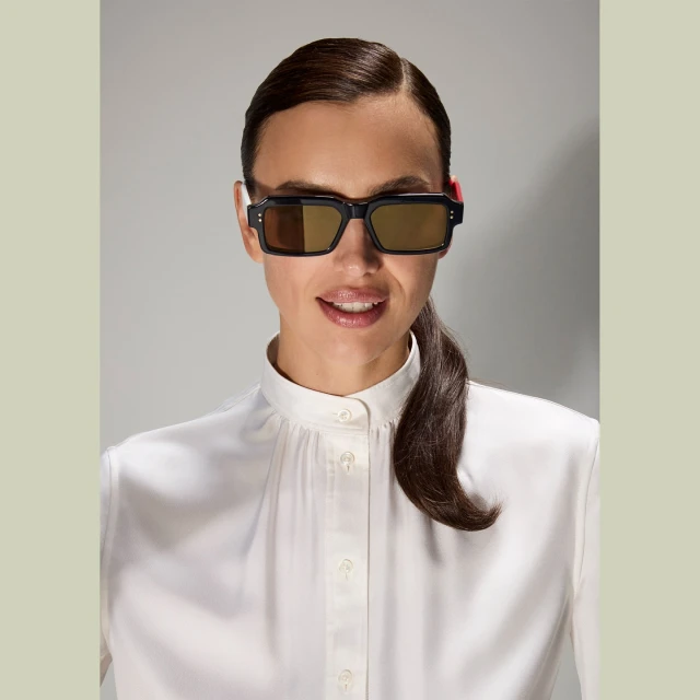 A woman in a white blouse and black sunglasses poses against a neutral background, showcasing fashion and beauty in a studio shot.