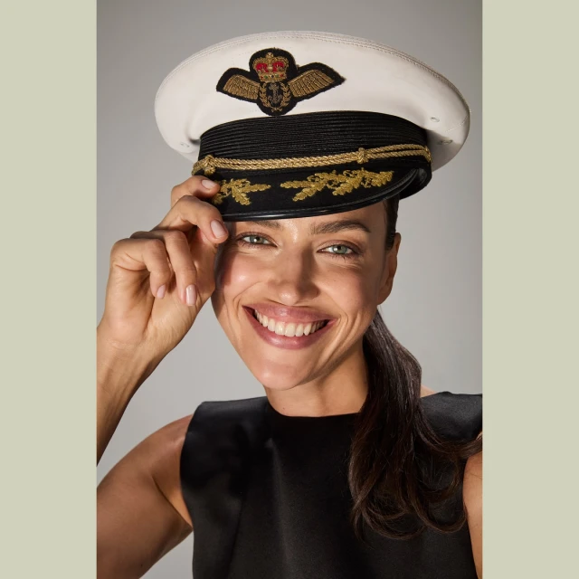 Smiling woman in a black dress wears a white naval hat with gold trim in a studio setting against a neutral background. Close-up shot.