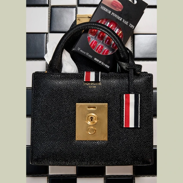 Black leather handbag with a gold clasp and nail tips on a black-and-white tiled surface, showcasing beauty and fashion accessories.