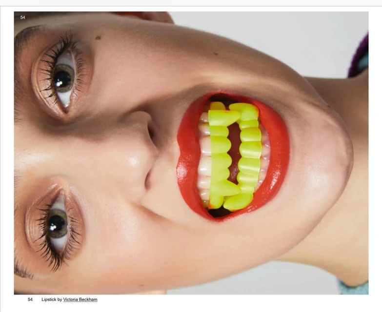 A close-up portrait captures a woman with red lipstick, eyes open, and a yellow plastic novelty teeth prop in her mouth, set against a light background.