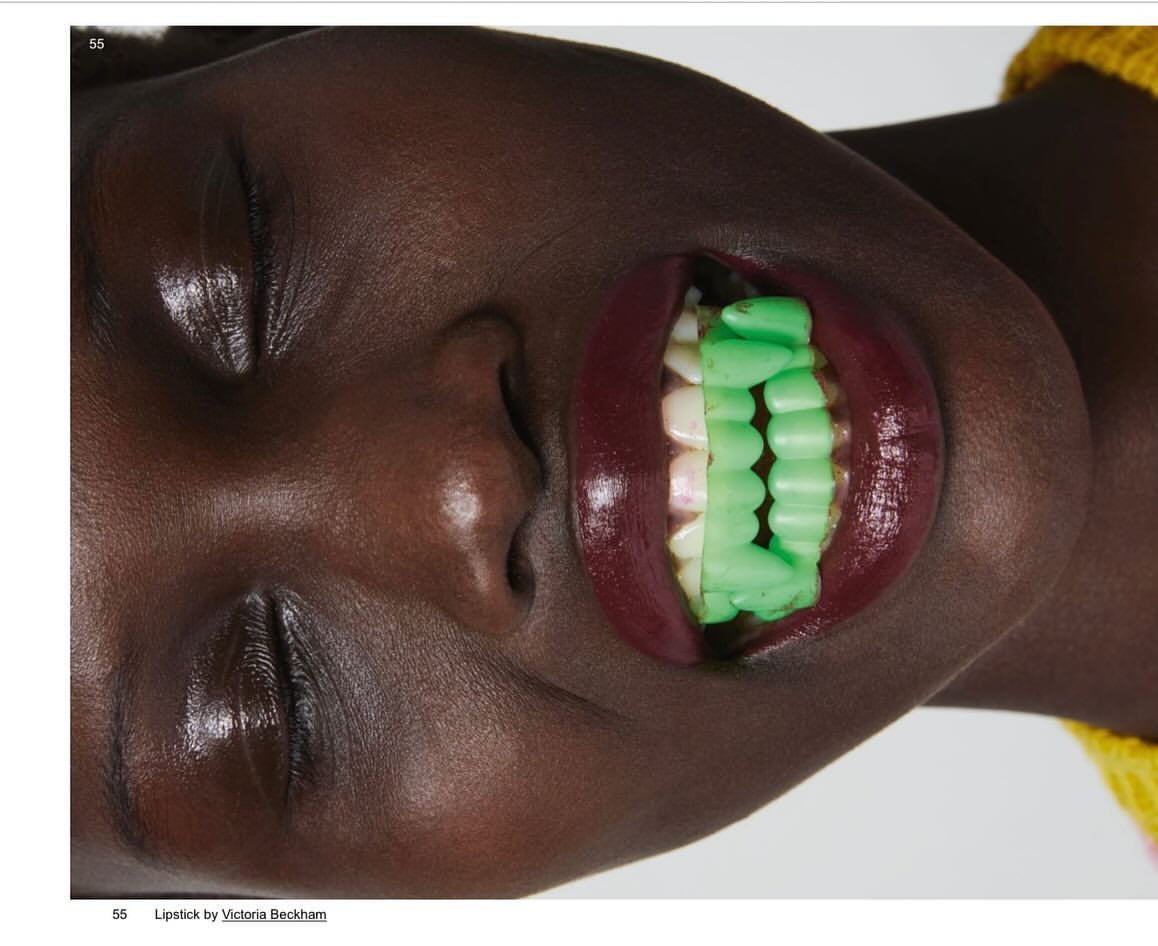 Close-up shot of an adult female with dark skin wearing red lipstick and green dental grills against a white background in a studio.