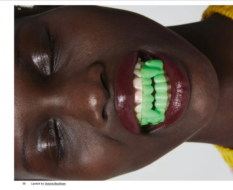 Close-up shot of an adult female with dark skin wearing red lipstick and green dental grills against a white background in a studio.