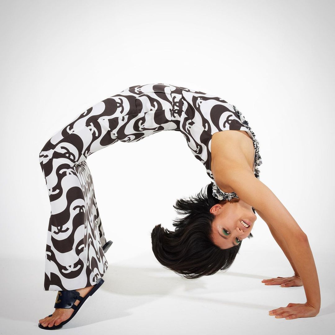 A brunette woman in a black and white patterned jumpsuit does a backbend on a white surface, showcasing fitness and flexibility.