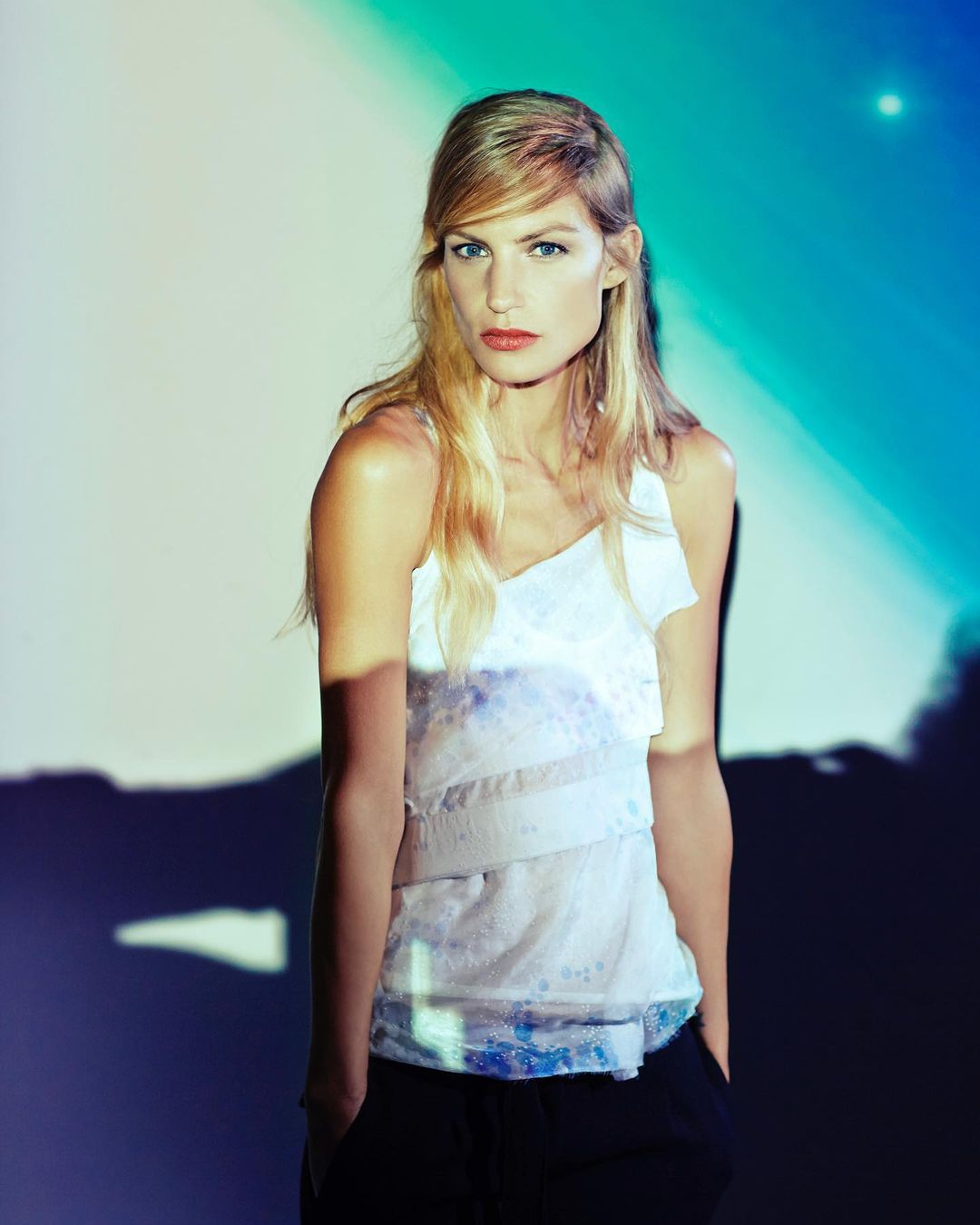 Portrait of an adult woman with fair skin, wearing a layered white top in front of a blue and white studio backdrop.
