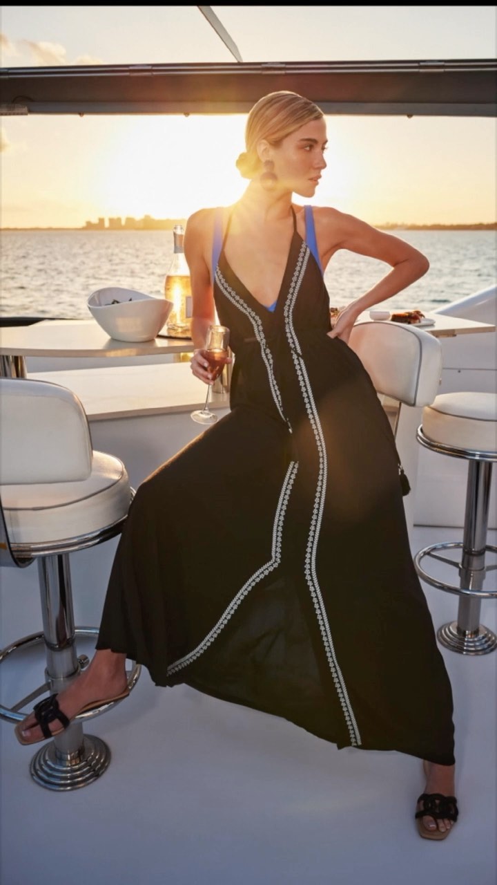 A woman in a black dress sits on a stool on a yacht deck at sunset. She holds a wine glass with a city skyline in the background.