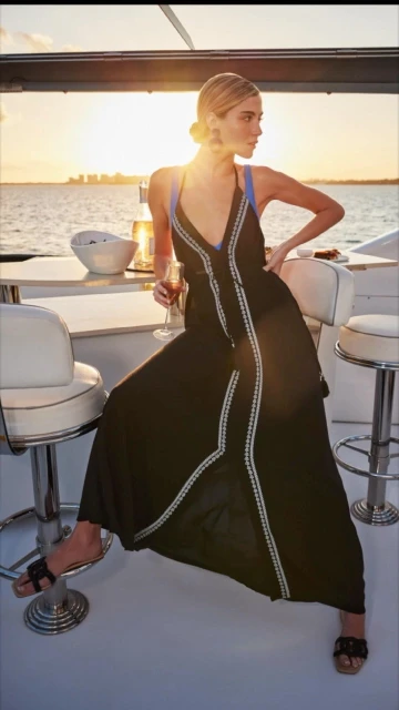 A woman in a black dress sits on a stool on a yacht deck at sunset. She holds a wine glass with a city skyline in the background.