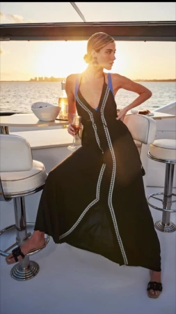 A woman in a black dress sits on a stool on a yacht deck at sunset. She holds a wine glass with a city skyline in the background.