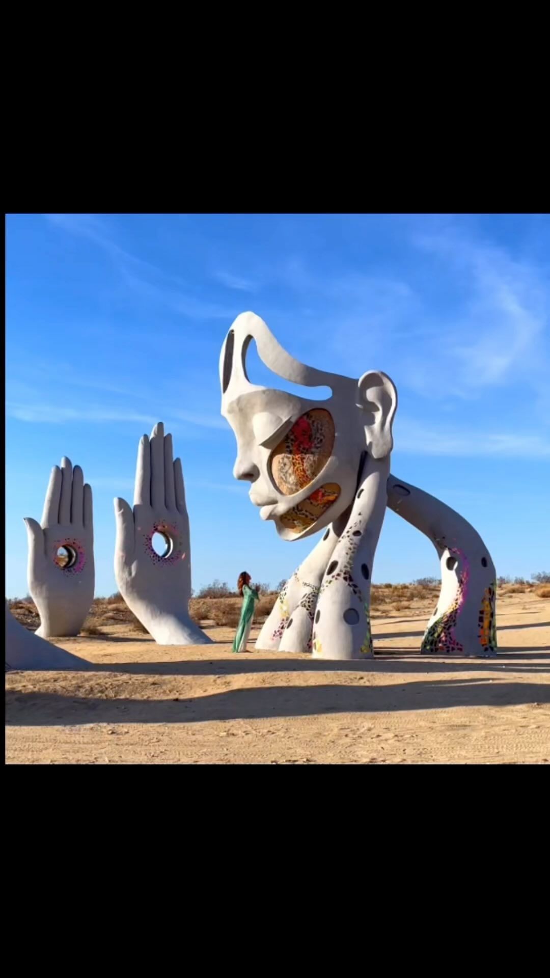 Sculpture of a face and hands in the desert with a woman standing nearby in a long dress, against a clear blue sky.