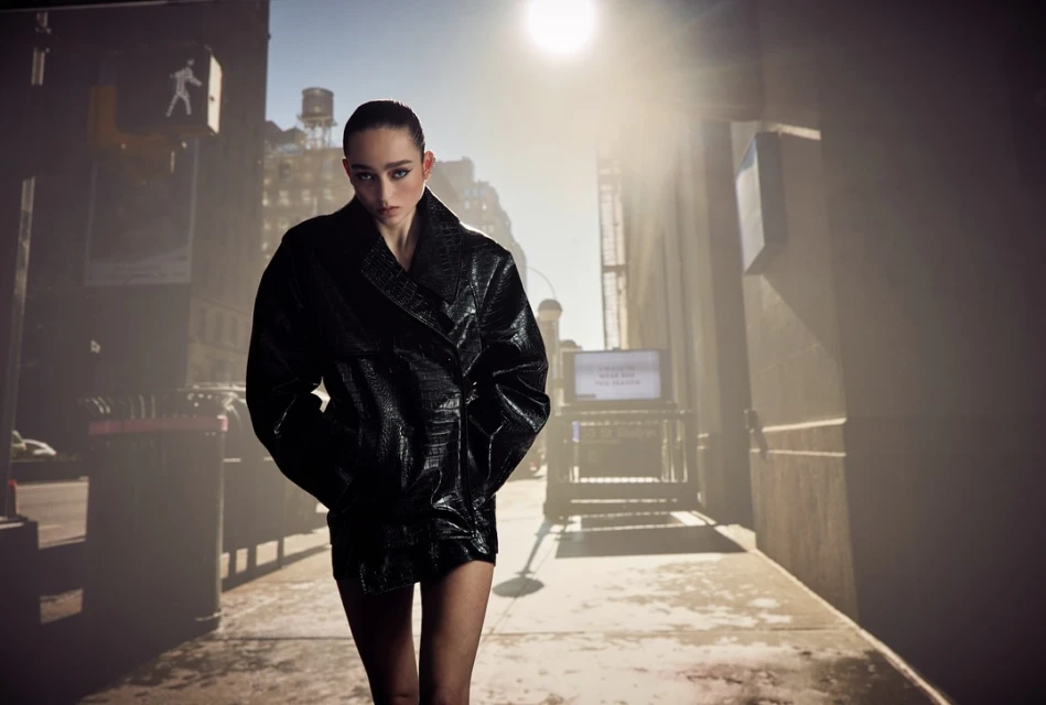 Fashionable woman in a dark coat stands in an urban setting with bright sunlight, creating a striking contrast and highlighting her attire.