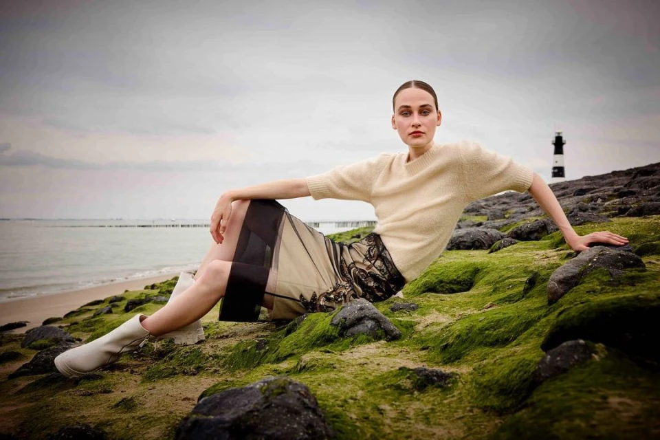 Woman in a beige sweater and a black lace skirt poses on a rocky, mossy surface near a body of water with a lighthouse in the distance.