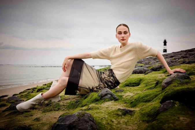 Woman in a beige sweater and a black lace skirt poses on a rocky, mossy surface near a body of water with a lighthouse in the distance.