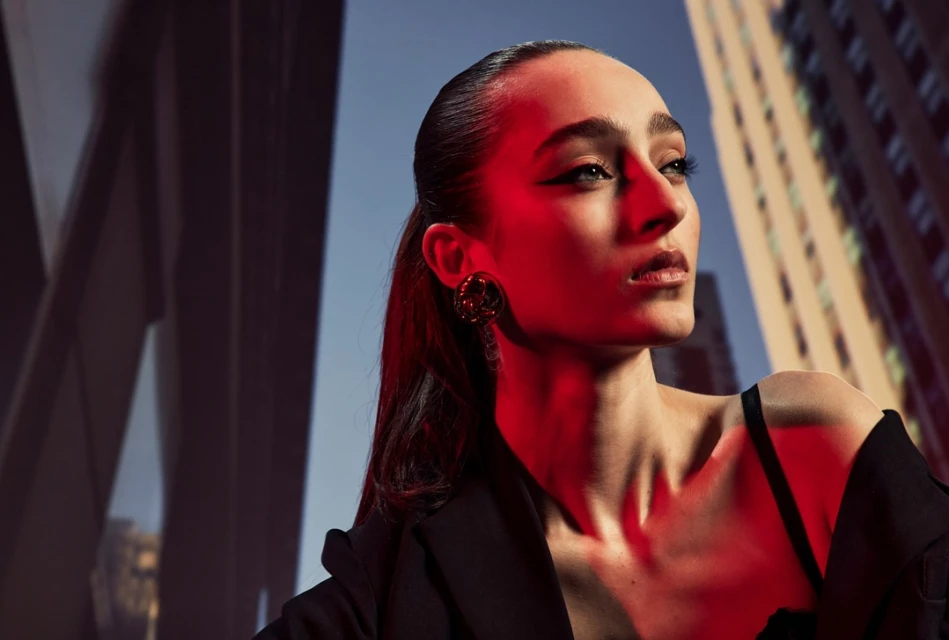 Portrait of a young woman with dark hair pulled back, wearing a black jacket and statement earrings, with an urban building background.