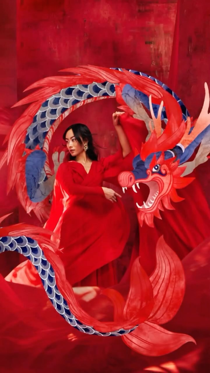 Elegant woman in a red dress posing next to a large, red and blue dragon sculpture in an indoor setting with draped red fabric.