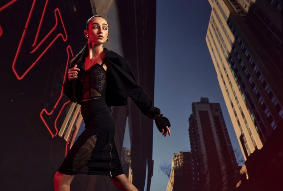 Woman in black lace top and skirt poses in front of a red neon light, with buildings in background, fashion photography, urban setting.