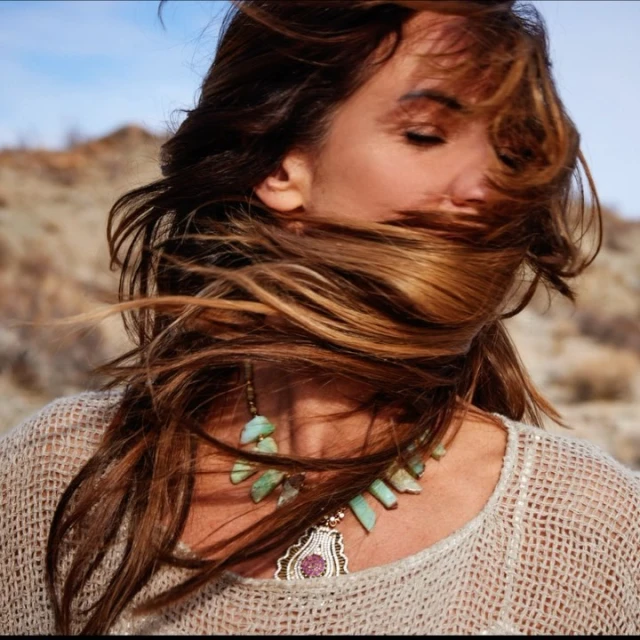 Adult woman in a desert landscape with windblown brown hair, wearing a knit top and a large stone necklace with tan and blue background.