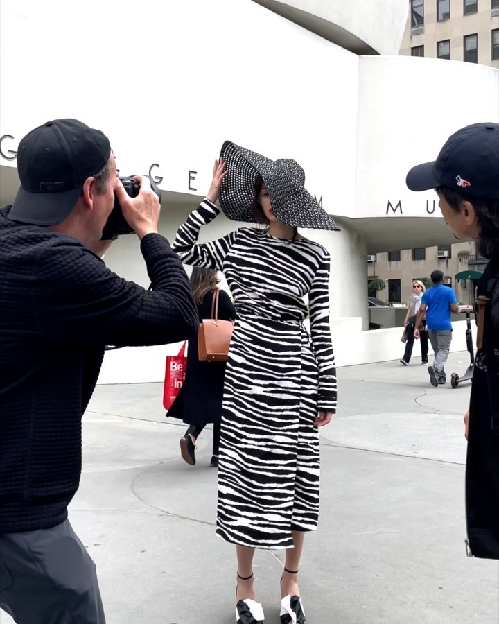 Fashion shoot in front of a museum featuring a woman in a zebra-print dress and large hat being photographed, with onlookers nearby.