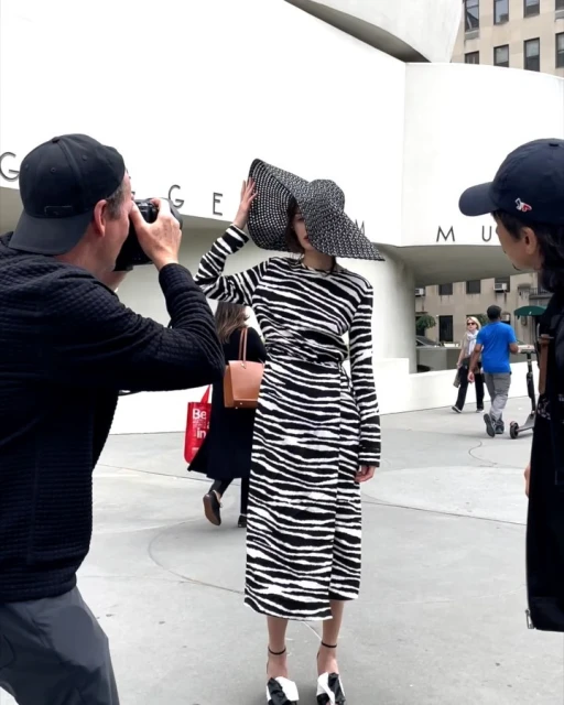 Fashion shoot in front of a museum featuring a woman in a zebra-print dress and large hat being photographed, with onlookers nearby.