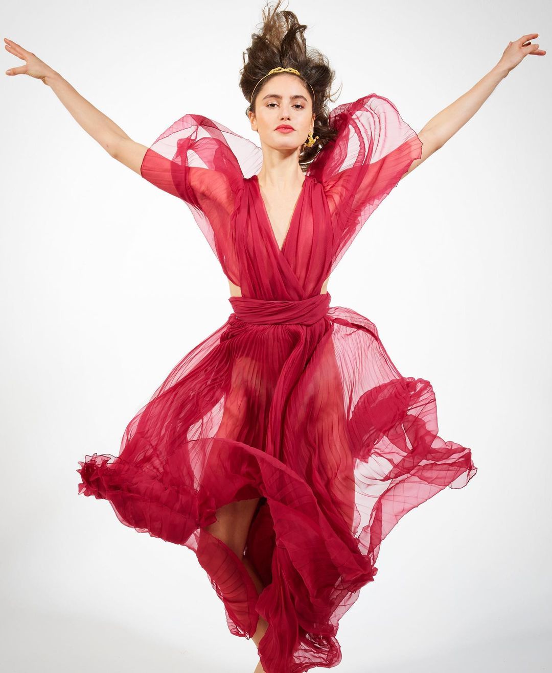 Elegant woman in motion wearing a flowing red dress with arms outstretched against a bright white background in a studio setting.