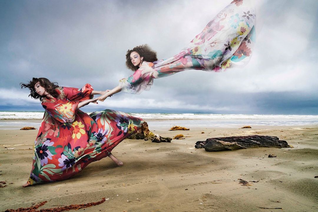 Two women on a beach holding hands, wearing colorful floral dresses that flow in the wind, with a cloudy sky and the ocean in the background.