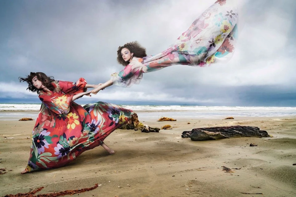 Two women on a beach holding hands, wearing colorful floral dresses that flow in the wind, with a cloudy sky and the ocean in the background.