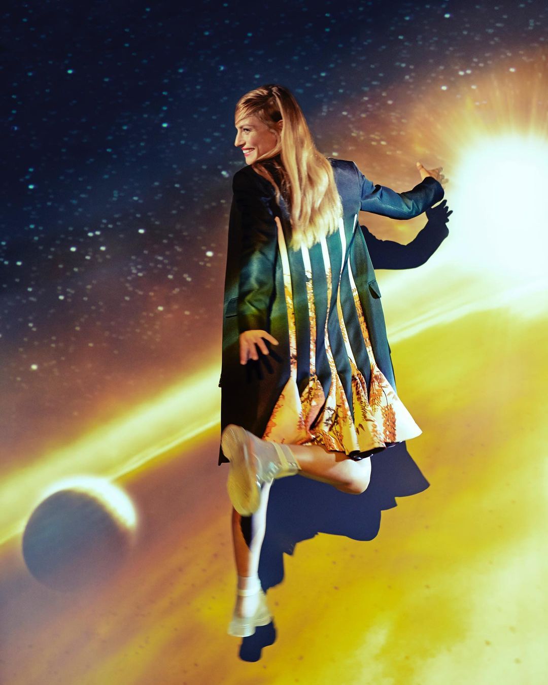 Blonde woman in a black and gold jacket poses with an outstretched hand against a background of planets and a bright sun.