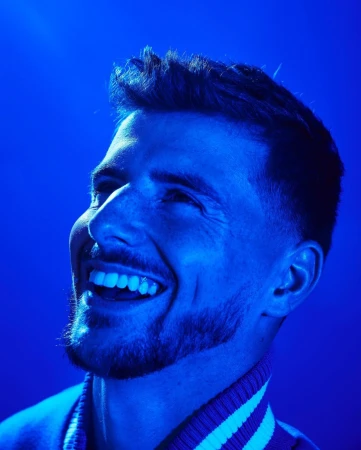 Portrait of a cheerful adult male with a beard, smiling and looking upwards, lit with vibrant blue lighting against a solid background.