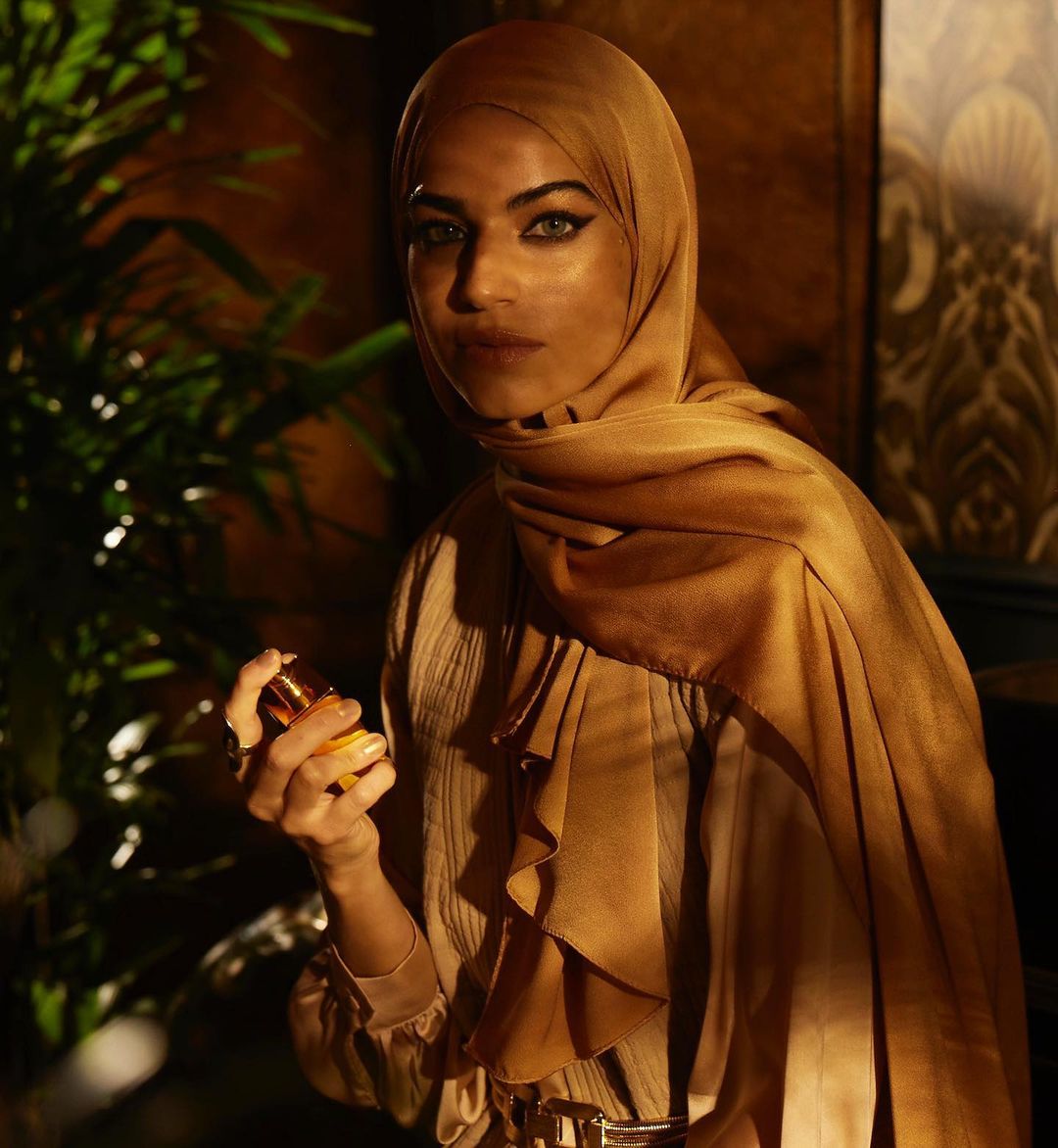 A woman in a hijab and formal attire holds a perfume bottle in front of a decorative background and green leafy plants.