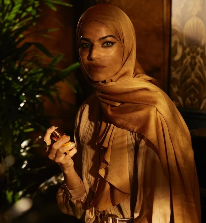 A woman in a hijab and formal attire holds a perfume bottle in front of a decorative background and green leafy plants.