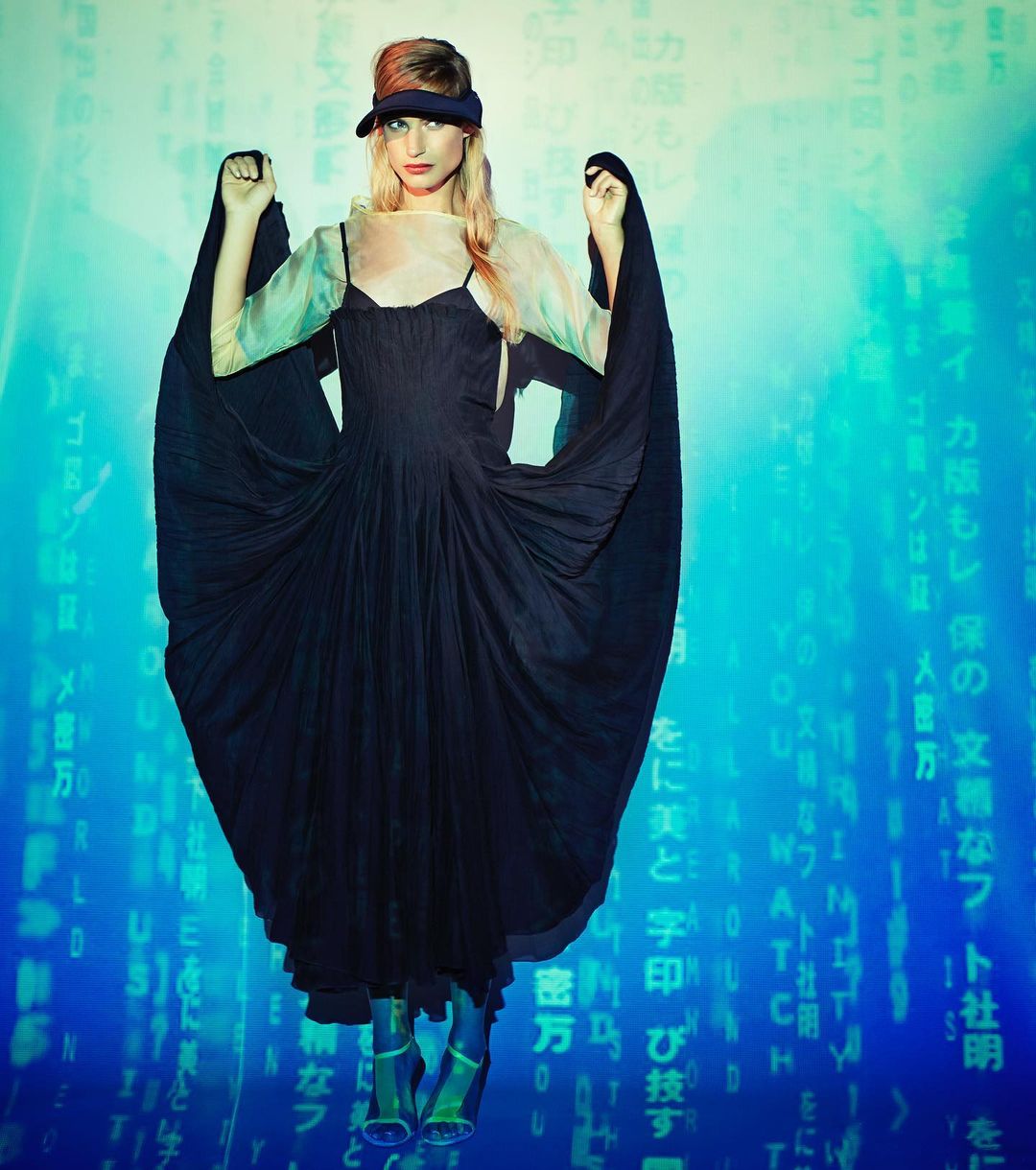 Woman poses in a black dress and visor against a matrix-style digital background. She wears semi-transparent boots in a studio setting.