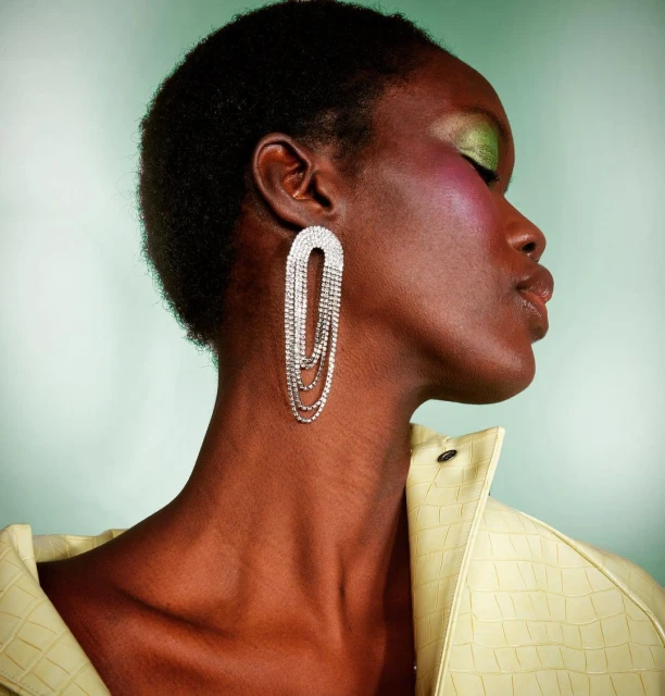 Profile portrait of a woman with dark hair wearing large, cascading rhinestone earrings and a pastel yellow jacket in a studio setting.